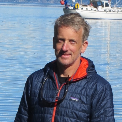 David Hand in a blue puffer jacket with a fishing boat in the background