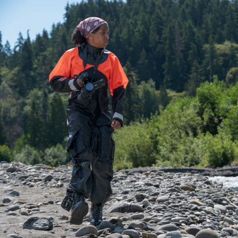 Person in black and orange wetsuit walks along rocky river shore on sunny day with forest in the background.