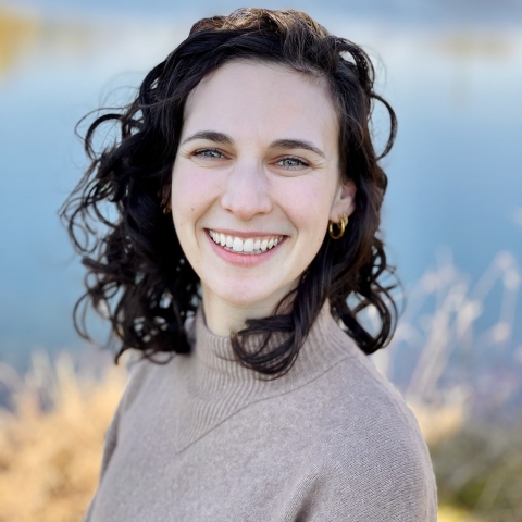 A woman with brown hair smiles into the camera in front of a body of water