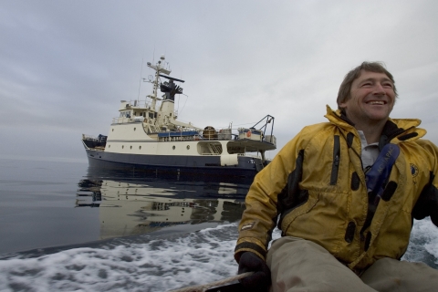 A man in yellow jacket drives a skiff away from a large yellow and blue ship.
