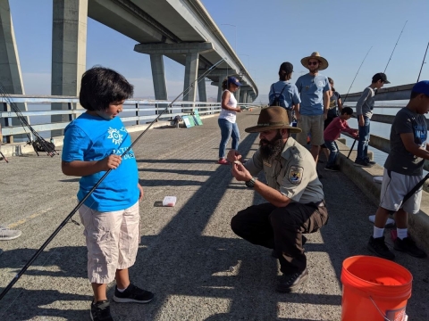 A man in a brown U.S. Fish and Wildlife Service uniform crouches down to help a boy bait his fishing rod hook