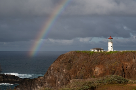 A rainbow arcs over the ocean and a lighthouse sitting on a rocky point.