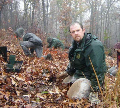 Mike Brod, a biologist with USFS, looks up as he sits amongst fallen leaves and plants smooth coneflower. There are other biologists doing the same in the background. Coats are wet and it appears to be raining. 