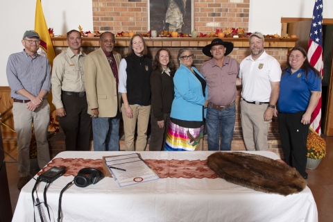 a group of nine people stand together smiling, behind a table with a large document and animal fur on it
