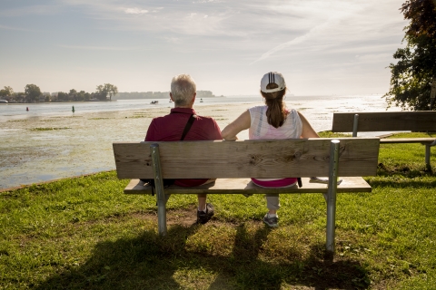 A man and a woman sit on a bench, with their backs to us, facing the river at Detroit River International Wildlife Refuge.