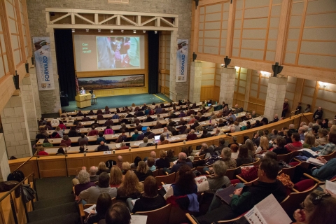 auditorium filled with people with large screen above the stage