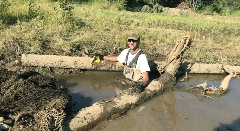 A grinning man in waders is up to his waist in muddy water, with muddy logs on either side of him.