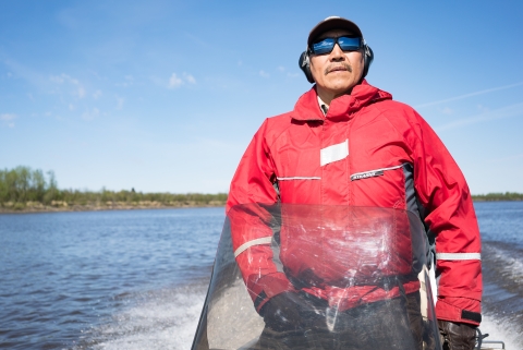 A man in an orange float coat drives a small skiff on a river