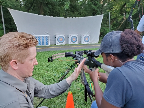 A man teaching a woman how to handle a crossbow while she aims at an archery target