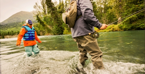 a low-angle shot of a man and a woman in fishing gear standing in rushing river water. The man is holding a fishing pole.