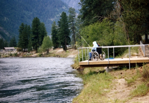 A man wearing a hoodie and sitting in a wheelchair fishes from a platform overhanging a river.