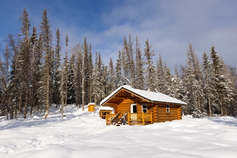 A wood cabin stands in the snow at the edge of a forest.