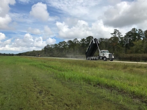 A dump truck tips a load of gravel onto a gravel road bordered in field and forest.