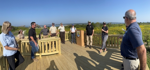 People stand in a circle on a wooden platform raised above green vegetation, with blue sky in the background.