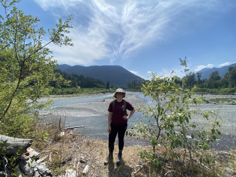 Service intern, Kathleen Gerard, poses in front of the Elwha River with mountains in the background.