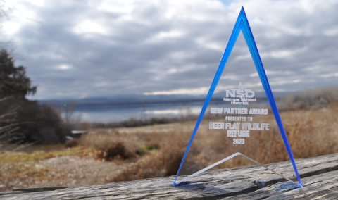 A translucent rectangular award sits in front of a lake and sagebrush habitat with an engraving that reads “Nampa School District New Partner Award Presented To Deer Flat National Wildlife Refuge 2023”