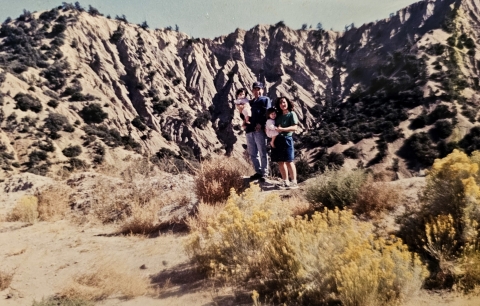 A couple stands holding two children in their arms in the center of the picture, with brush in front of them and cliff faces in the background.