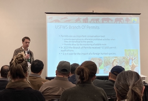A person stands in front of seated people, holding a microphone, and next to a projector screen that has the title, "USFWS Branch of Permits"