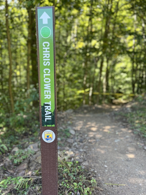 An image of a trail marker that reads "Chris Clower Trail." In the background is a trail that leads through the forest.