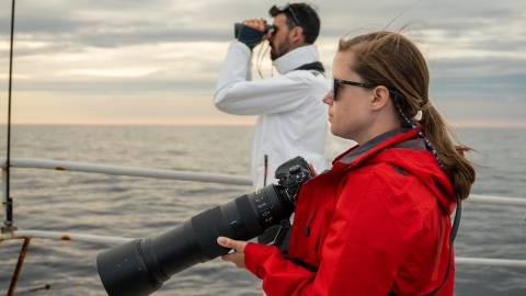 A woman wearing sunglasses and a red jacket standing in the foreground holding a camera with a large lens and looks out at sea on a ship. In the background, a man in a white jacket stands looking through a pair of binoculars.
