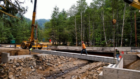 Construction equipment and staff at work on concrete structures spanning over top of a creek bed.