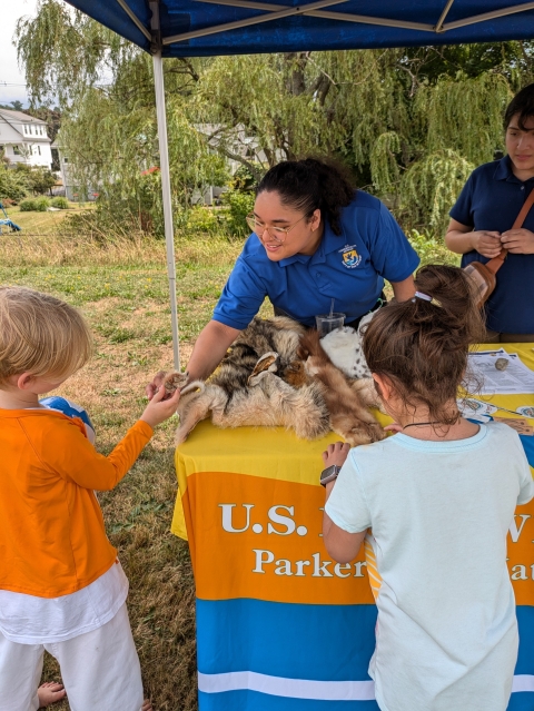 a woman in a blue USFWs uniform smiles while teaching kids about wildlife at an informational table outside