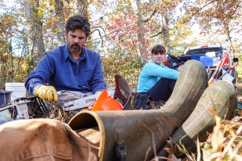 People seated on the ground, one with a chainsaw bar and a small tool in hand
