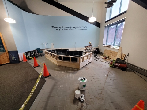 A large room showing the floor which is partly tile and partly carpeted. Sitting on the floor also are orange cones with hazard tape connecting the cones. This is being done to keep visitors out of a construction area. Withing construction area a platform is being built. 