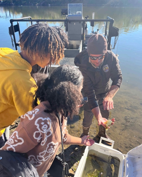 Image of uniformed USFWS biologist showing two students a fish in front of a boat,