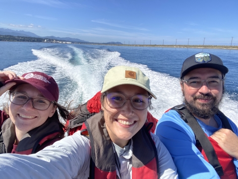 Three service members sit grouped together on the back of a boat. Water with a wake can be seen in the background.