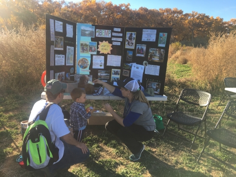 A staff member teaches a child and their guardian in front of a trifold presentation in a field
