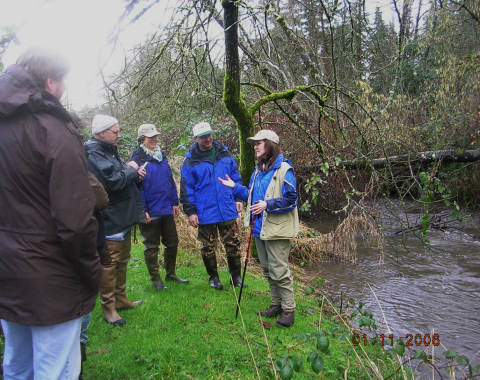 A group of people stand in the grass along a creek. A woman speaks with a walking stick in her hand as the others listen. 
