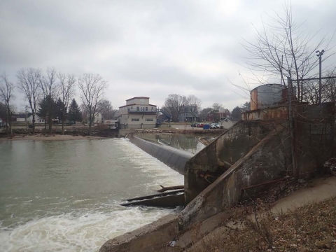 A concrete dam on a river under an overcast sky. Water flows over the dam into the lower river. Leafless trees and bare branches are visible, along with a few buildings and a fenced area nearby. The scene is quiet and wintry.