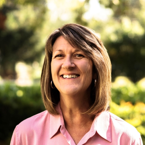 A woman with brown hair and wearing a pink, collared shirt smiles brightly