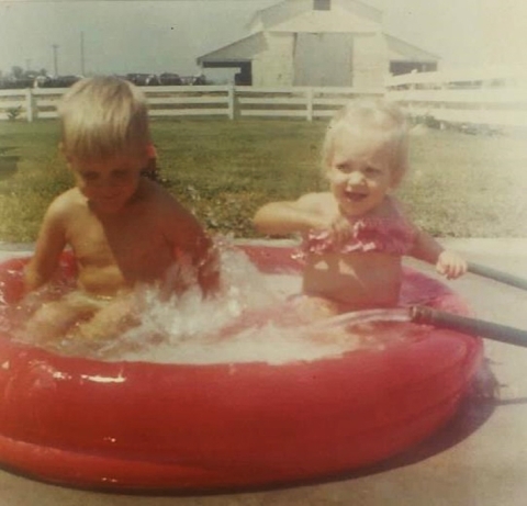 An old, washed out photo of two small, happy children in a small, red plastic pool full of water. A hose is filling the pool