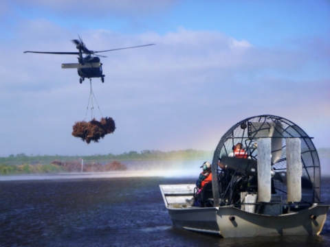 a helicopter carries trees over a water body and near an air boat