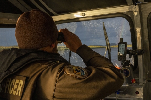 a uniformed officer in a covered helm of a boat looks through binoculars through the windshield to another section of a river. 