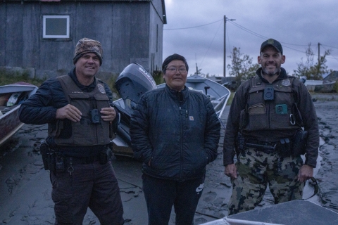 two uniformed officers pose for a picture with a village patrol officer between them on the muddy riverbank with boats grounded in the background. 