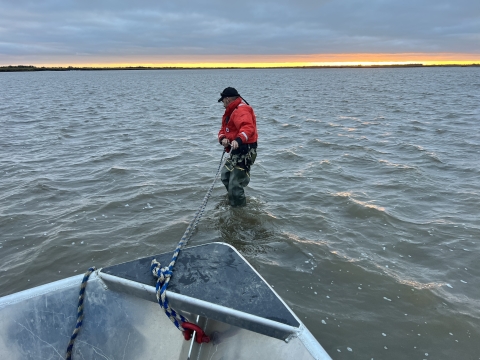 a uniformed officer in a large float coat stands in the middle of a large river puling a rope tied to the bow of a boat. 