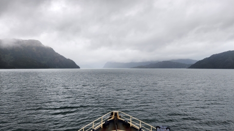 Bow of a boat looking across the ocean with foggy mountains on the horizon