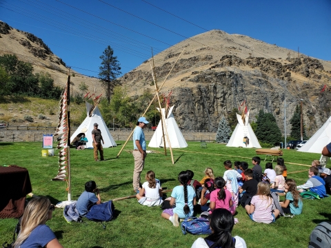 Students sit in a grass field facing 4 tipis and two indigenous educators