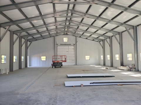 Vast interior space of an all metal building. Windows line the walls at person height, The framing is thick steel. There is a scissor lift and some excess panels in the image