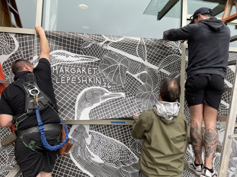 Three people work together to apply an adhesive panel featuring birds to window at the entrance of a buildling