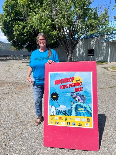 A woman in a blue shirt stands next to a red sign reading "Winthrop Kids Fishing Day"