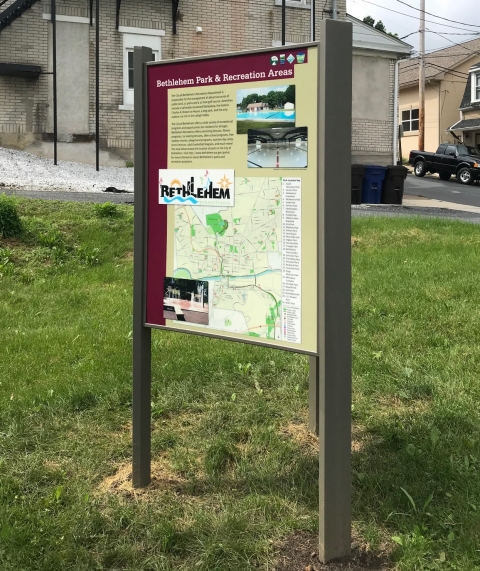 a sign with text and a map stands in green grass, with a brick building in the background
