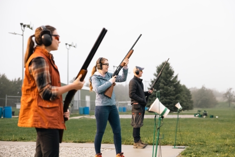 Three people wearing eye and ear protection stand with firearms at a skeet target range.
