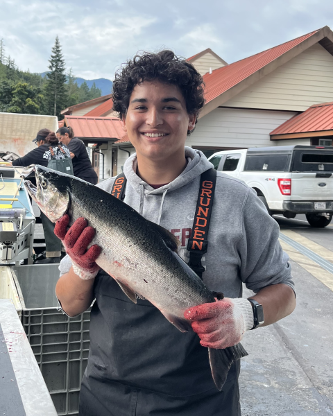 AmeriCorps Service Member holding an adult coho salmon at Quilcene National Fish Hatchery. Hatchery vehicle and building in the background.
