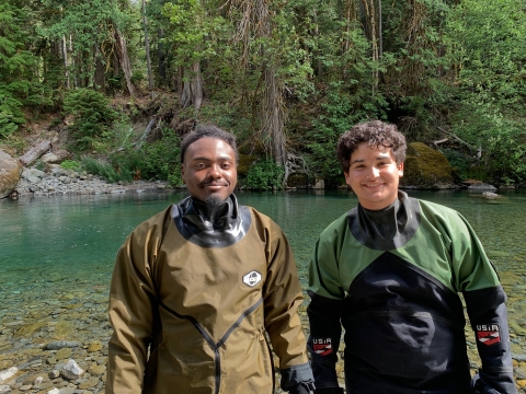 Two field crew members standing in dry suits on bank of river. River and trees in the background.