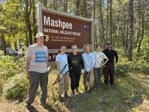 6 people volunteers stand in front of Mashpee NWR entrance sign.