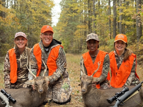 Four women hunters in camo and orange sit in a forest with a shot deer.